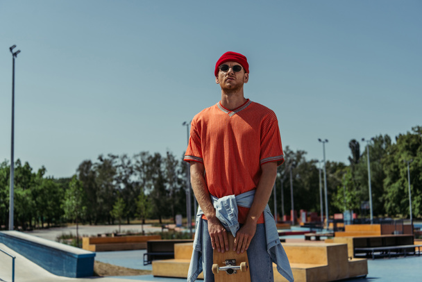 модный человек со скейтбордом стоит в скейт-парке в солнцезащитных очках и красной шапочке - Фото, изображение