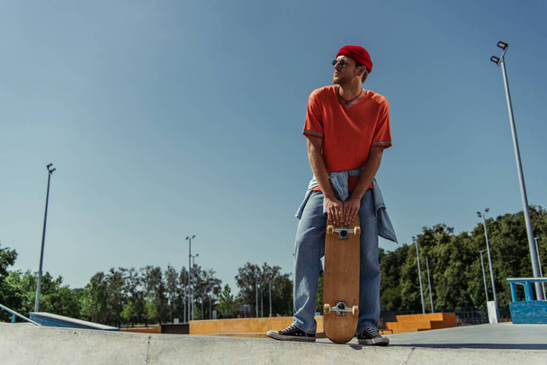 full length of man in orange t-shirt and jeans standing with skateboard in park - Fotó, kép