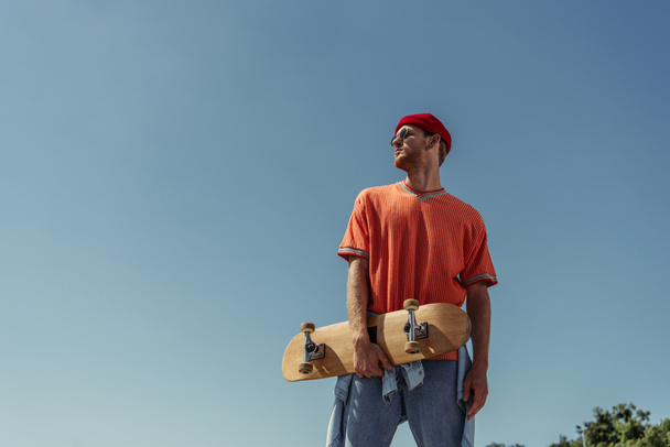 low angle view of man in sunglasses and beanie standing with skate under blue sky - Fotó, kép