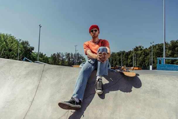full length of stylish skateboarder in gumshoes sitting on ramp in skate park - Foto, afbeelding