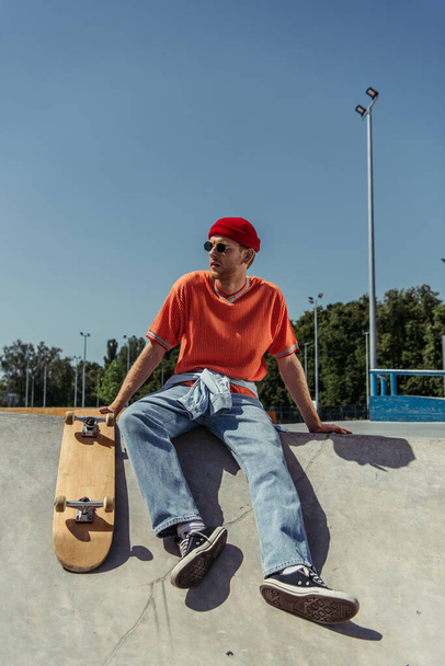 full length of fashionable man in gumshoes and sunglasses sitting on ramp near skate - Foto, Bild