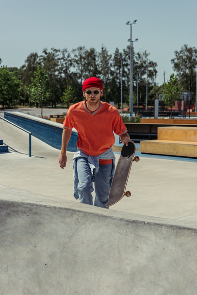 young man in red beanie and sunglasses walking on ramp with skateboard - Fotografie, Obrázek