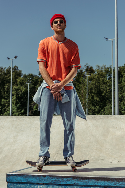 full length of young man in jeans and sunglasses standing on skate in park - Photo, Image