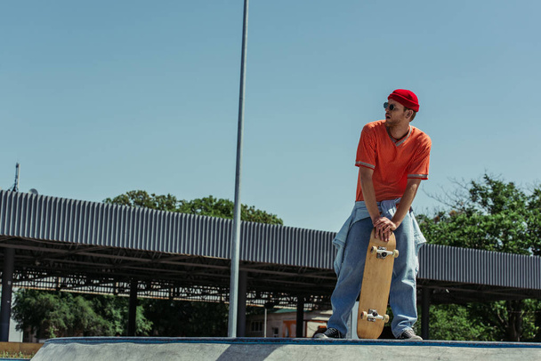 Модный мужчина в полный рост с скейтбордом, отводящим взгляд на улицу - Фото, изображение