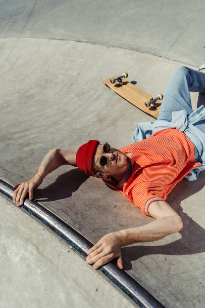 high angle view of man in sunglasses and beanie looking at camera while lying on ramp near skate - Photo, Image