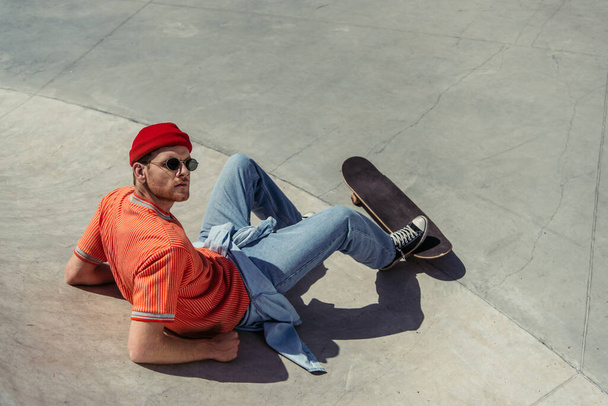 high angle view of man in sunglasses and trendy clothes sitting on ramp near skateboard - Foto, Imagen