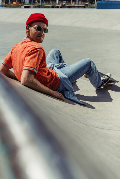 fashionable skateboarder looking at camera while sitting on ramp on blurred foreground - Zdjęcie, obraz