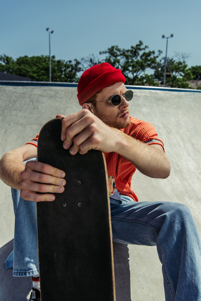 young and trendy man in sunglasses sitting with skateboard and looking away - Foto, afbeelding