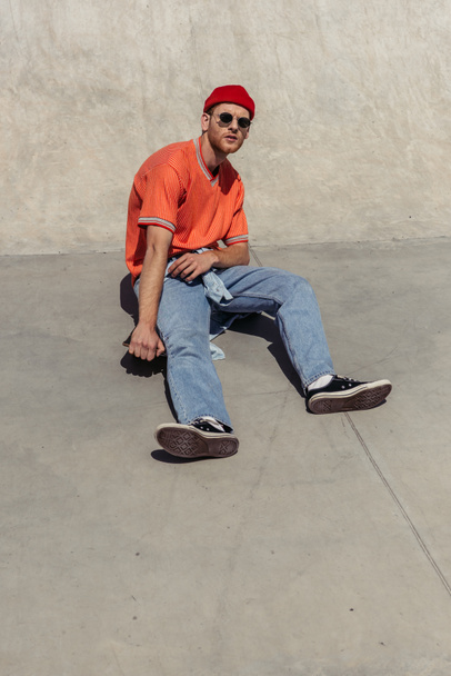 full length of man in sunglasses and jeans sitting on skate in park - Foto, imagen