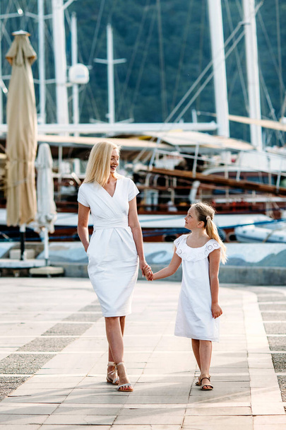 Happy middle aged millenial mother and blonde teenager daughter in white dresses walking by the harbor and marina with yachts in a touristic sea resort with sailboats and mountains on background - Fotó, kép
