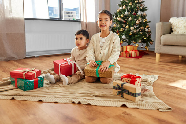 christmas, winter holidays and childhood concept - happy little girl and boy with gifts sitting on floor in front of each other at home - Foto, Imagem