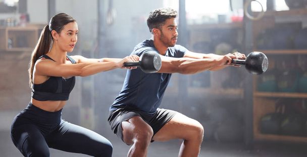 Slim, fit and gym couple exercising with dumbbell equipment, doing lower body core workout or training in a wellness facility. Young woman and man doing squatting exercise for perfect form or health. - 写真・画像