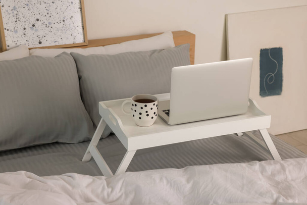White tray table with laptop and cup of drink on bed indoors - Fotoğraf, Görsel