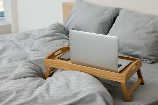 Wooden tray table with laptop and smartphone on bed indoors - Photo, image