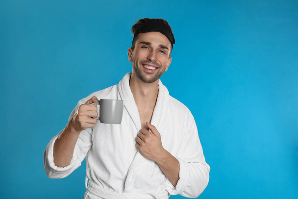 Young man in bathrobe with cup of coffee on light blue background - Фото, зображення