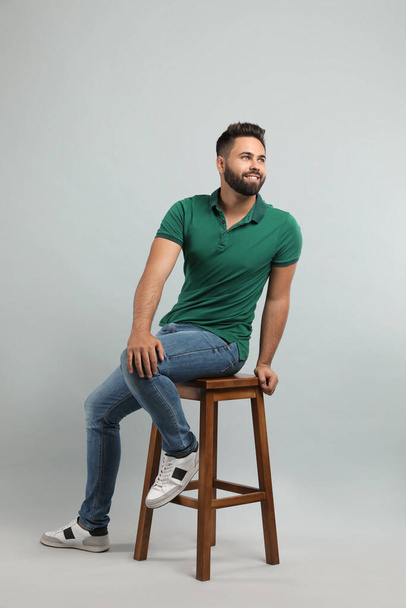 Handsome young man sitting on stool against light grey background - Фото, изображение