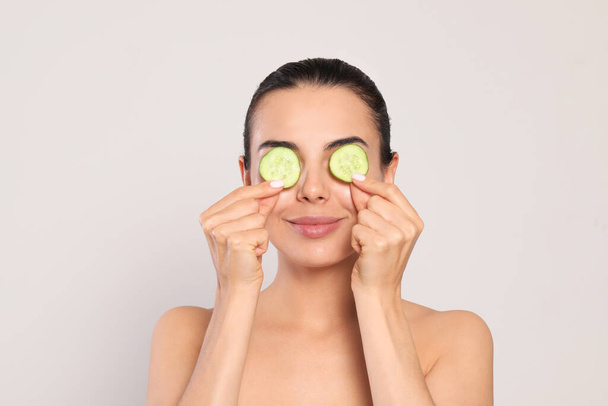 Beautiful young woman putting slices of cucumber on eyes against light grey background - Fotoğraf, Görsel