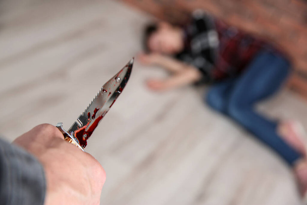 Man with bloody knife and his victim on floor indoors, closeup. Dangerous criminal - Foto, afbeelding