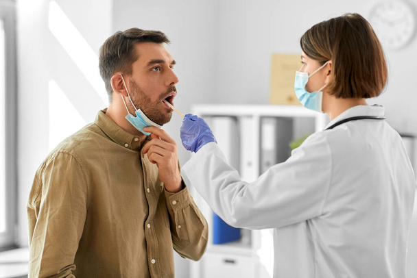 medicine, healthcare and people concept - female doctor in mask with tongue depressor checking sore throat of man patient at hospital - Foto, Imagen