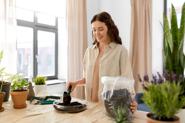 people, gardening and housework concept - happy woman with trowel and soil in bag planting flowers in glass vase at home - Foto, Bild