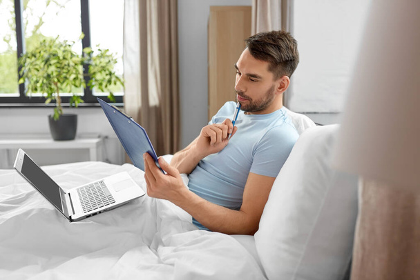 people, technology and remote job concept - man with laptop computer and clipboard working in bed at home bedroom - Φωτογραφία, εικόνα