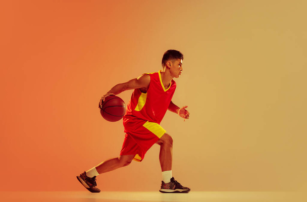Portrait of young man, basketball player training isolated over orange studio background in neon light. Concentration. Concept of healthy lifestyle, professional sport, hobby, power and strength - Foto, imagen