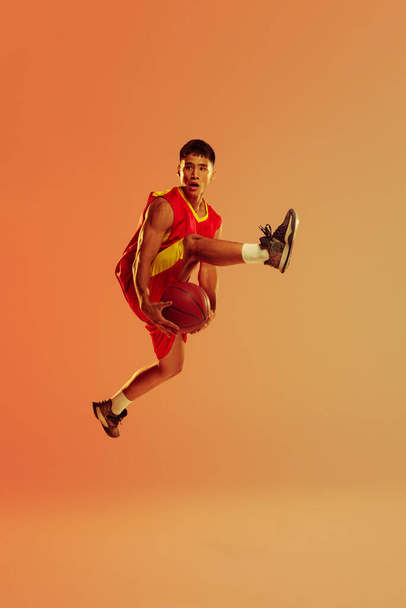 Portrait of young man, basketball player in motion, jumping isolated over orange studio background in neon light. Concept of healthy lifestyle, professional sport, hobby, power and strength - Fotoğraf, Görsel