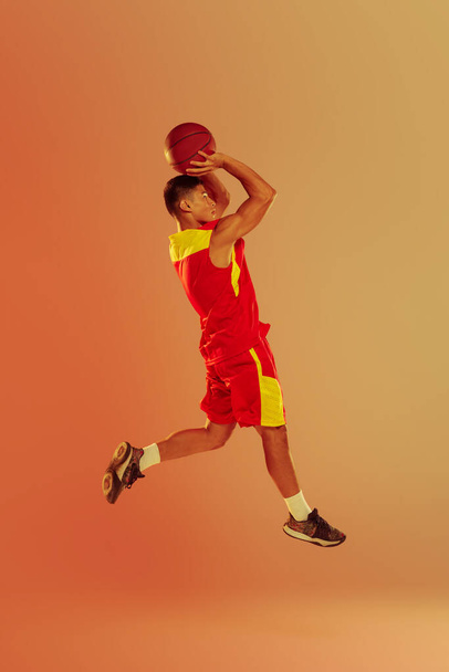 Portrait of young man, professional basketball player training isolated over orange studio background in neon light. Jump shot. Concept of healthy lifestyle, professional sport, hobby, power, strength - Photo, image