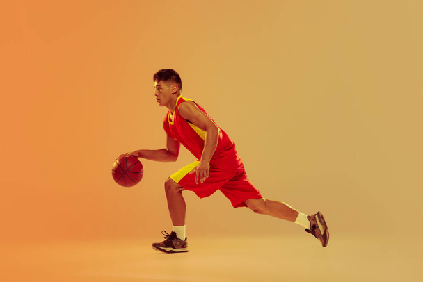 Πορτρέτο του νεαρού, προπόνηση μπασκετμπολίστα, dribbling απομονωμένη πάνω από πορτοκαλί φόντο στούντιο σε νέον φως. Έννοια του υγιεινού τρόπου ζωής, του επαγγελματικού αθλητισμού, χόμπι, δύναμη και δύναμη - Φωτογραφία, εικόνα