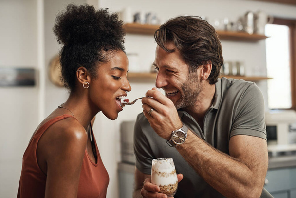 Romantic, happy and interracial couple eating a healthy yogurt together in a cute, sweet and fun kitchen romance. Loving, in love and excited husband feeding his beautiful afro wife delicious dessert. - Foto, imagen