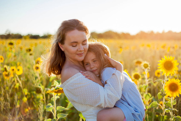 Mom hugs her little daughter. A beautiful young woman with a 4-year-old child in her arms in a sunflower field. - Foto, imagen