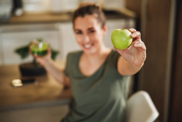 Shot of a smiling woman holding a green apple and glass of smoothie. Selective focus on an apple. - Fotografie, Obrázek