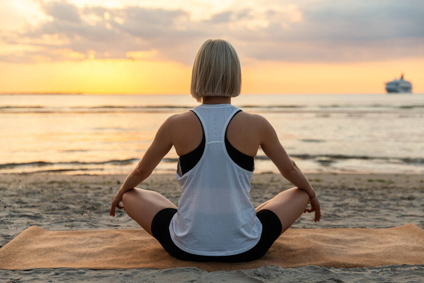 yoga, mindfulness and meditation concept - woman meditating in lotus pose on beach over sunset - 写真・画像