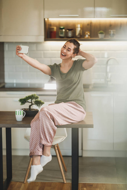 Молода щаслива жінка робить селфі зі своїм смартфоном, п'ючи ранкову каву на своїй кухні вдома
. - Фото, зображення