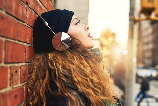 Music is my escape. a young woman listening to music while leaning against a brick wall outside - Photo, Image