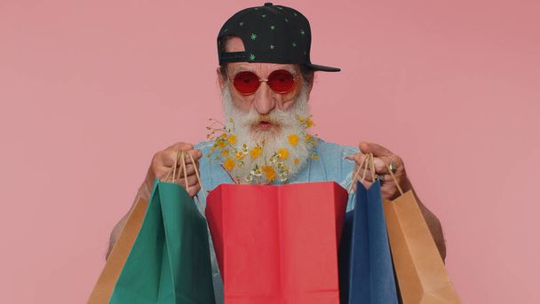 Senior man with flowers in gray-haired beard showing shopping bags, advertising discounts, smiling looking amazed with low prices, shopping on Black Friday holidays. Elderly grandfather isolated alone - Fotografie, Obrázek