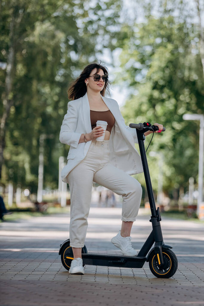 Beautiful young woman in white suit and sunglasses standing with her electric scooter in city parkland and drinking coffee - Photo, Image