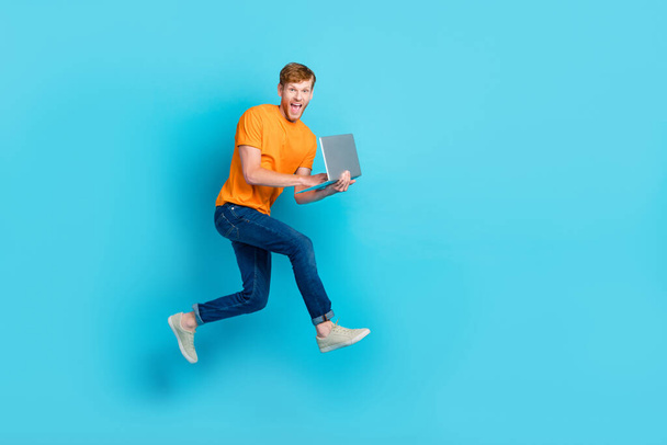Full size profile photo of hooray red hair guy run with laptop wear t-shirt jeans boots isolated on blue color background. - Zdjęcie, obraz