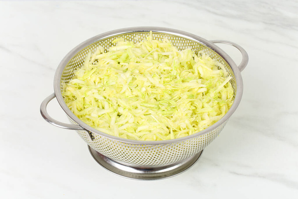 Grated salted zucchini placed in a steel microperforated colander, for straining out the water of them, over marble white kitchen board. Preparing marrow vegetables for freezing or for cooking - Photo, image