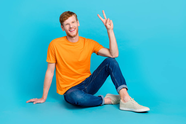 Full length photo of cheerful funny guy dressed orange t-shirt sitting floor showing v-sign isolated turquoise color background. - Foto, Bild
