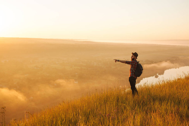 Side view of young bearded male traveler in casual clothes and hat with backpack, pointing away while standing on grassy slope of mountain during hiking trip at sunrise - Foto, afbeelding