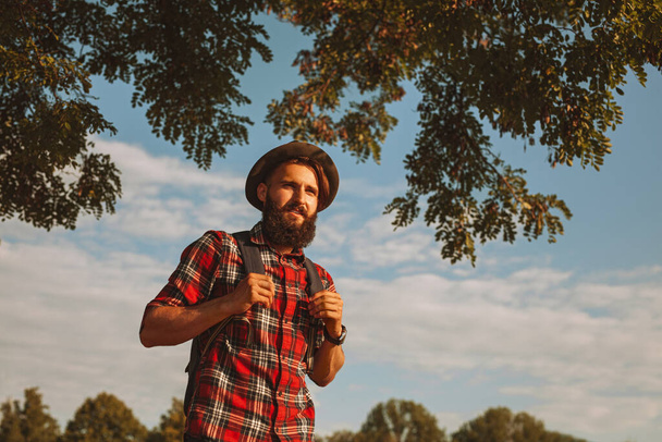 From below bearded man in checkered shirt with hat and backpack walking under lush tree branches, against cloudy blue sky while exploring countryside in weekend - Foto, imagen
