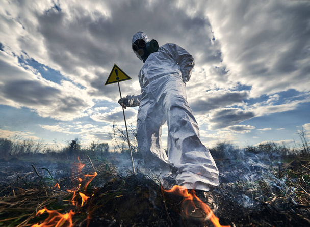 Πυροσβέστης οικολόγος καταπολέμηση φωτιά στο πεδίο. Άνδρας με προστατευτική στολή ακτινοβολίας και μάσκα αερίου κοντά σε φλεγόμενο γρασίδι με καπνό, κρατώντας προειδοποιητικό σήμα με θαυμαστικό. Έννοια φυσικής καταστροφής. - Φωτογραφία, εικόνα