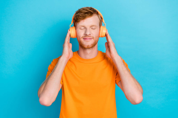 Photo of attractive guy wear orange outfit arms touch headphones closed eyes enjoying music isolated on cyan color background. - Фото, изображение