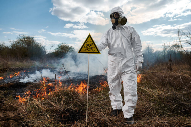 Fireman ecologist fighting fire in field. Man in protective radiation suit and gas mask near burning grass with smoke, holding yellow triangle with skull and crossbones warning sign. - Foto, Imagen