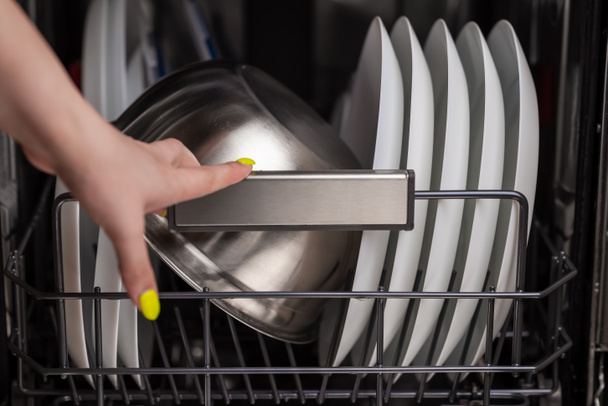 Close-up of a dishwasher basket with clean white plates and a metal bowl, a woman's hand pulls out clean dishes. Small depth of field. - Zdjęcie, obraz