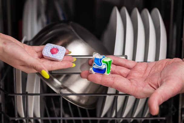 Close-up of young woman and man hands holding two colored capsule for the dishwasher. In the background, out of focus, is a dishwasher with clean dishes. Small depth of field. - Photo, Image