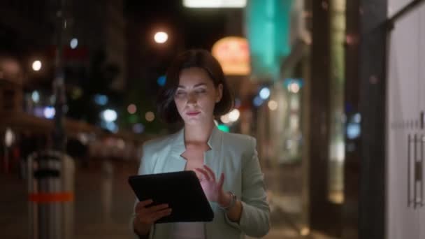 Καυκάσιος έξυπνο μοντέρνο εστιασμένο θηλυκό tablet περιήγησης, ενώ το περπάτημα στο κέντρο της πόλης το βράδυ. Αυτοπεποίθηση κυρία κουβέντα με τους συναδέλφους σε απευθείας σύνδεση μετά την εργασία - Πλάνα, βίντεο