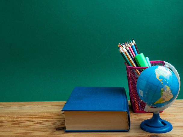 A book, a metal cup with pencils and a globe on the table against the background of a green board. School. - Photo, Image