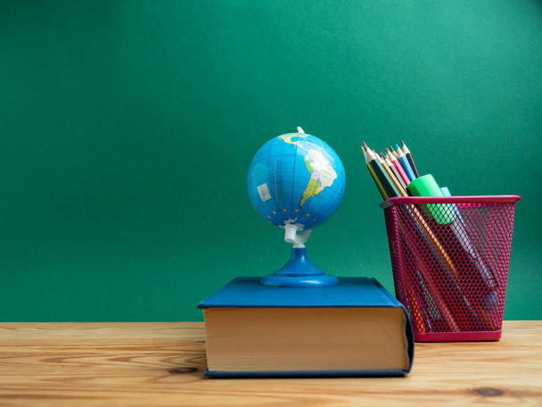 Ένα βιβλίο, ένα μεταλλικό κύπελλο με μολύβια και μια σφαίρα στο τραπέζι με φόντο ένα πράσινο πίνακα. Σχολείο. - Φωτογραφία, εικόνα
